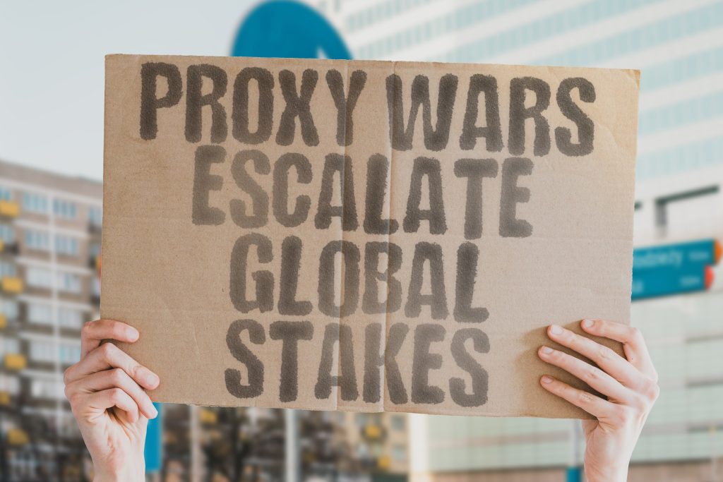 Cardboard sign with the message “Proxy Wars Escalate Global Stakes” held at a protest, symbolizing global tensions and international risks.
