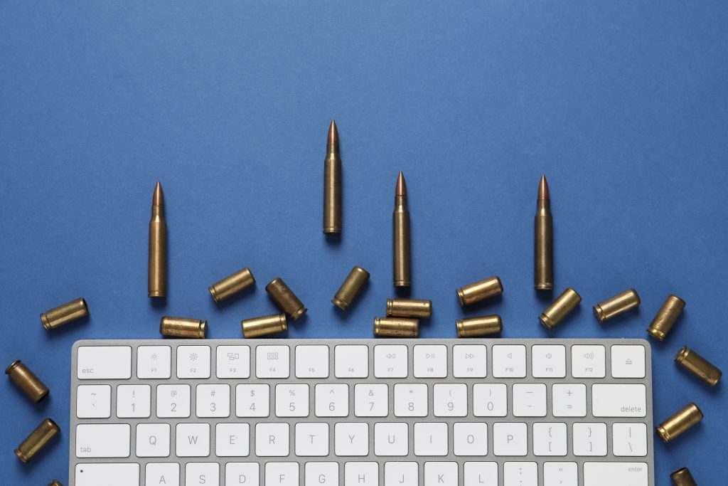 A computer keyboard surrounded by bullets on a blue background, illustrating hybrid warfare, cyber conflict and the intersection of digital and physical threats.