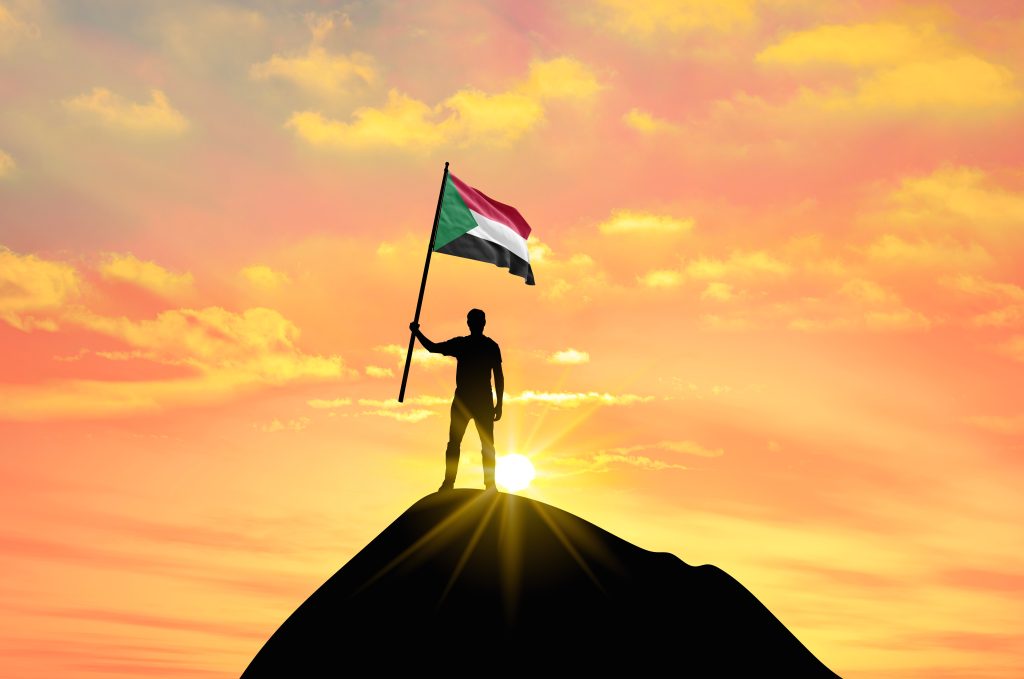 A silhouette holding the Sudanese flag at sunrise symbolises hope, resilience, and the demand for civilian leadership amid Sudan’s ongoing conflict.
