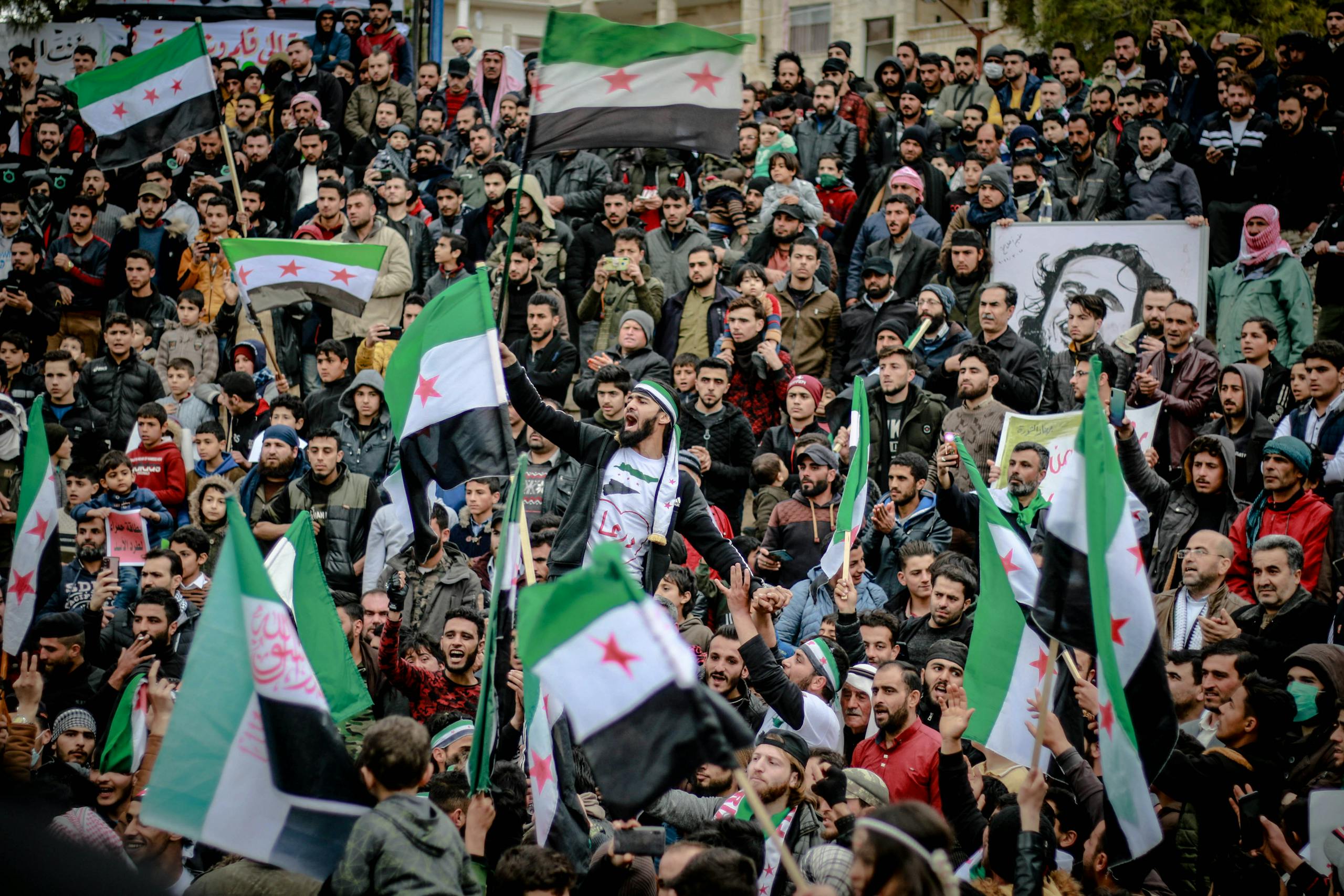 A large crowd gathers in a city square waving Syrian flags during a protest.