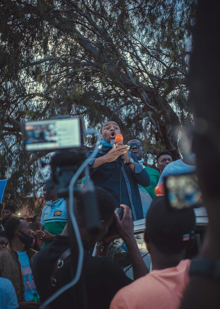 A speaker addresses a crowd with passion, captured through various recording devices outdoors.