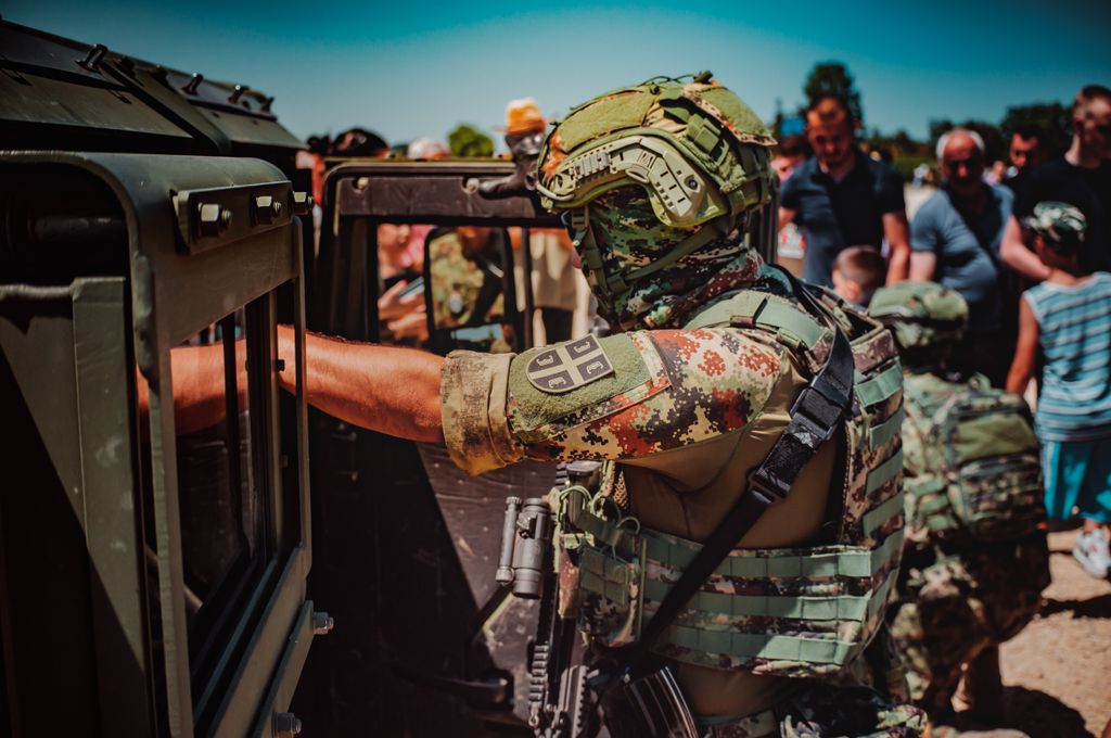 Military personnel surrounding an army vehicle in a public gathering on a sunny day.