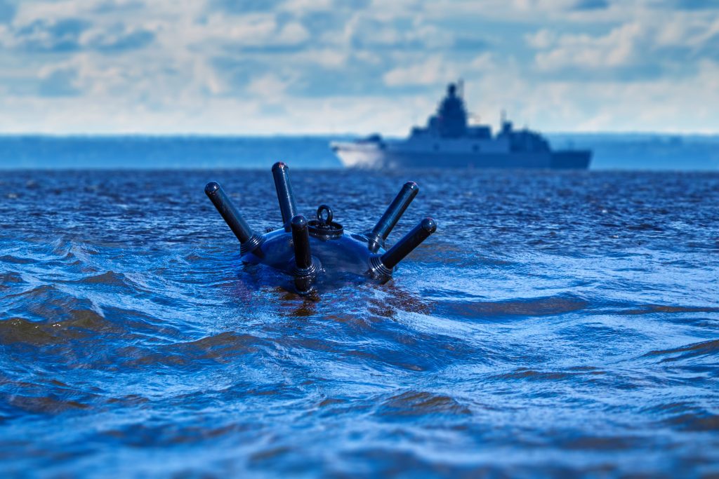 Naval sea mine in ocean with warship in distance illustrating maritime conflict, shipping disruption, and strategic chokepoint risk